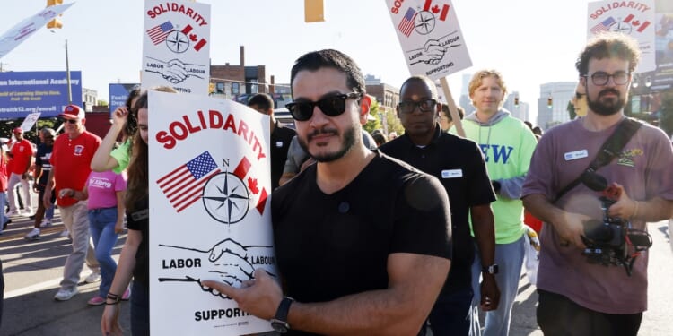 A group of people march holding Solidarity signs showing U.S. and Canadian flags, a compass, and shaking hands. The crowd appears diverse and determined, participating in a labor support event on a city street.