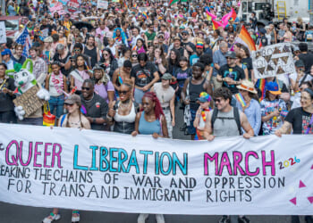 Transgender activists at a march. Pictured: a diverse crowd marches, holding a banner reading Queer Liberation March: Breaking the Chains of War and Oppression for Trans and Immigrant Rights amid colorful signs and pride flags.