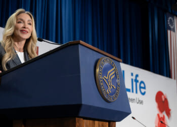 Dr. Stephanie Haridopolos in business attire speaks at a podium with the Department of Health and Human Services seal. A large sign behind her reads Life. Blue curtains and an American flag are visible in the background.