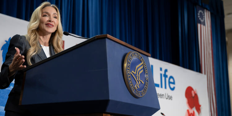 Dr. Stephanie Haridopolos in business attire speaks at a podium with the Department of Health and Human Services seal. A large sign behind her reads Life. Blue curtains and an American flag are visible in the background.