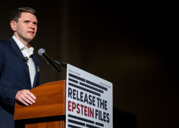 A man in a suit speaks at a podium with a sign reading RELEASE THE EPSTEIN FILES in bold letters, next to a microphone, against a dark background.