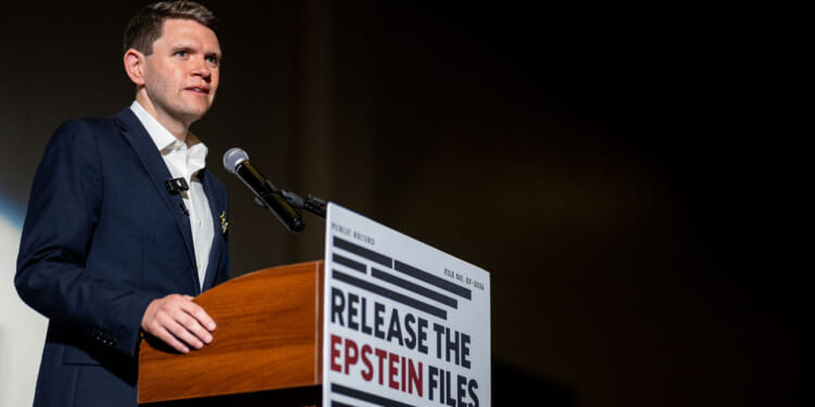 A man in a suit speaks at a podium with a sign reading RELEASE THE EPSTEIN FILES in bold letters, next to a microphone, against a dark background.