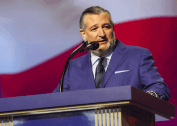 Sen. Ted Cruz in a blue suit speaks at a podium with a microphone, with a large red, white, and blue background behind him.
