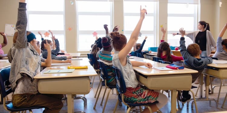 Kids in a classroom seated at their desks are raising their hands.