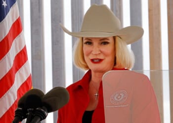 A woman in a tan cowboy hat and red shirt speaks at a podium with microphones, standing beside a U.S. flag and vertical blinds in the background.
