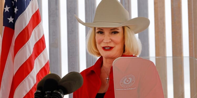 A woman in a tan cowboy hat and red shirt speaks at a podium with microphones, standing beside a U.S. flag and vertical blinds in the background.