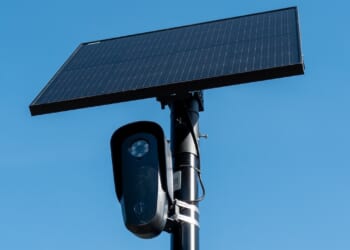 A black security camera is mounted on a pole beneath a large rectangular solar panel against a clear blue sky.