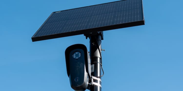 A black security camera is mounted on a pole beneath a large rectangular solar panel against a clear blue sky.