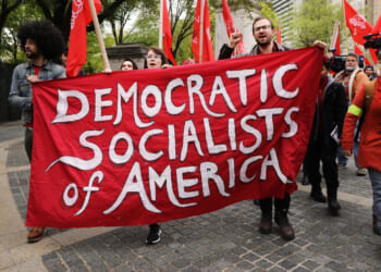 A group of people march outdoors holding a large red banner that reads Democratic Socialists of America. Some hold flags and raise fists; trees and buildings are visible in the background.
