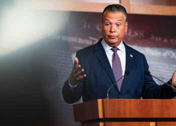 Alex Padilla in a suit and tie stands at a podium with microphones, gesturing with his hands while speaking. The background is dark with indistinct text.