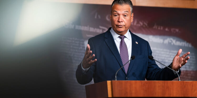 Alex Padilla in a suit and tie stands at a podium with microphones, gesturing with his hands while speaking. The background is dark with indistinct text.