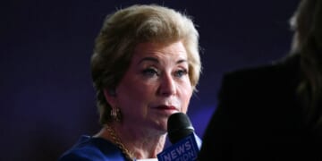 A woman with short blonde hair and gold earrings holds a NewsNation microphone, speaking during an interview. The background is dark and another person is partially visible in the foreground.