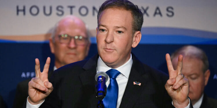 A man in a suit and blue tie stands at a podium, holding up both hands making peace signs, with two men blurred in the background and a banner reading Houston, Texas behind him.