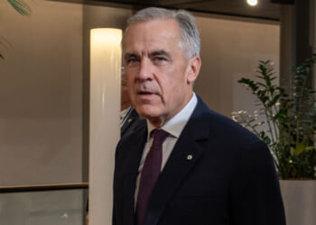 Mark Carney in a suit and tie stands indoors near plants and a light fixture, looking slightly to the side with a neutral expression, reminiscent of Mark Carney’s composed demeanor.