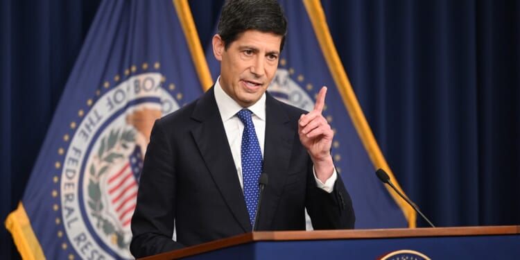 A man in a suit speaks at a podium with the Federal Reserve seal, raising one finger, with blue flags displaying the seal in the background.