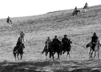 A group of U.S. special forces ride horses across a sloping, grassy landscape, embodying a quiet sense of heroism. Some riders are in the foreground while others are spaced out further back. The image is in black and white.