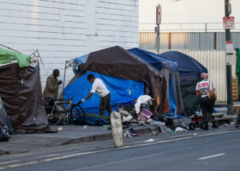 Several people interact near makeshift tents and belongings on a city sidewalk, with scattered items and bicycles, depicting an urban homeless encampment along a street—an unsettling scene that evokes images of modern dark ages within the city’s heart.