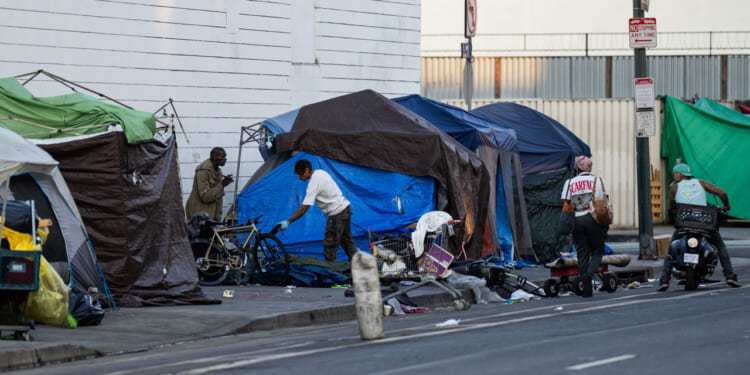 Several people interact near makeshift tents and belongings on a city sidewalk, with scattered items and bicycles, depicting an urban homeless encampment along a street—an unsettling scene that evokes images of modern dark ages within the city’s heart.
