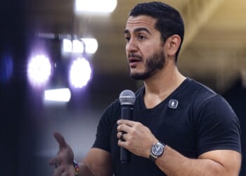 A man with dark hair and beard speaks into a microphone, wearing a black shirt and watch, gesturing with his left hand. Bright lights and a blurred background suggest he is at an indoor event or conference.