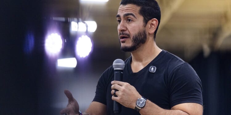 A man with dark hair and beard speaks into a microphone, wearing a black shirt and watch, gesturing with his left hand. Bright lights and a blurred background suggest he is at an indoor event or conference.