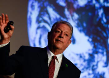 Al Gore in a suit speaks while holding a remote, with a large image of Earth projected behind him.