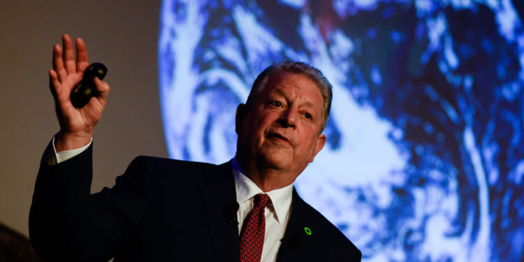 Al Gore in a suit speaks while holding a remote, with a large image of Earth projected behind him.