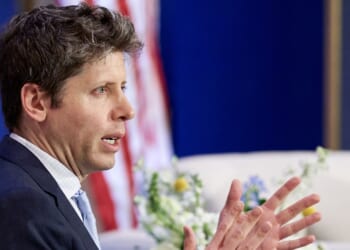 A man in a suit speaks and gestures with his hands, perhaps discussing the impact of AI. There is an American flag and some flowers in the background.