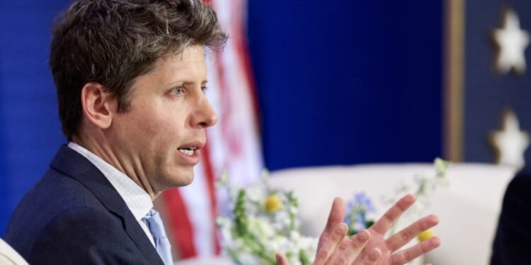 A man in a suit speaks and gestures with his hands, perhaps discussing the impact of AI. There is an American flag and some flowers in the background.