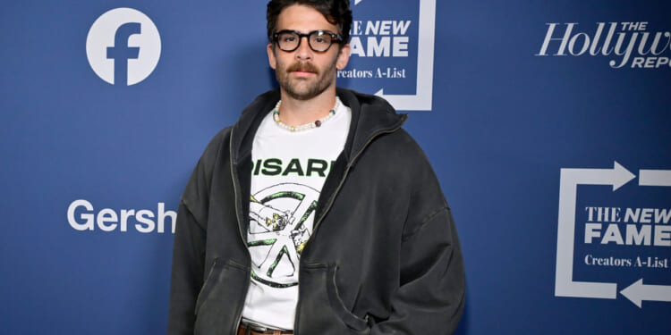 A man with dark hair, glasses, and a mustache stands in front of a blue backdrop with logos. He wears a white graphic t-shirt, a dark hoodie, and jeans.
