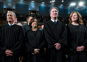 Four judges in black robes stand together in a large, ornate hall with an audience seated and standing behind them. The hall has blue patterned walls, bright lighting, and high ceilings.