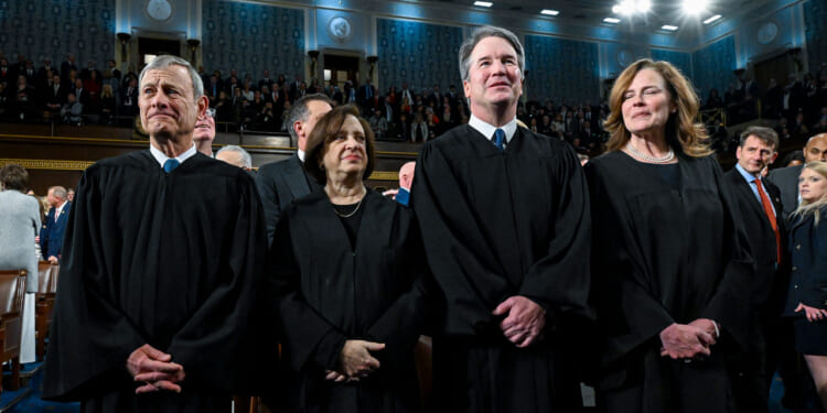Four judges in black robes stand together in a large, ornate hall with an audience seated and standing behind them. The hall has blue patterned walls, bright lighting, and high ceilings.