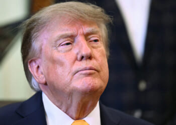 Donald Trump in a suit and yellow tie looks slightly upward with a serious expression, standing indoors with a blurred background.