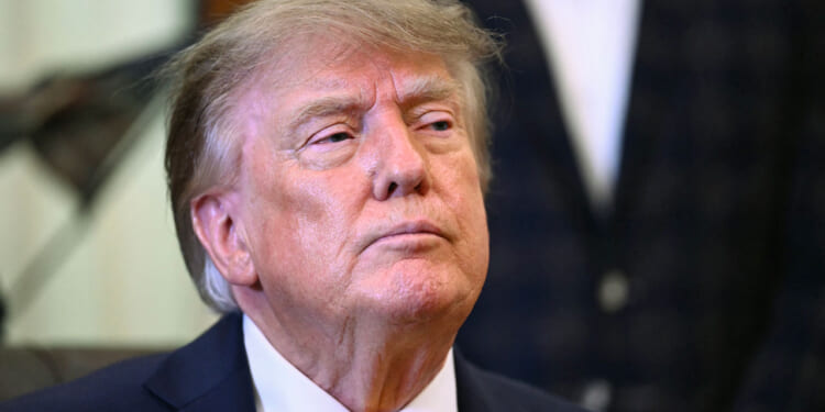 Donald Trump in a suit and yellow tie looks slightly upward with a serious expression, standing indoors with a blurred background.