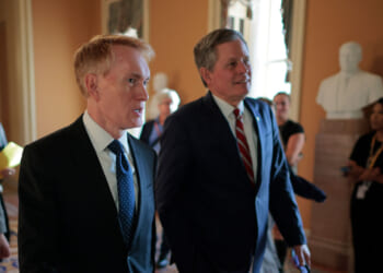 Sen. James Lankford and Sen. Steve Daines in suits walk through a hallway with others in the background. A white statue is visible on a pedestal, and sunlight streams through windows, casting a warm glow on the scene.