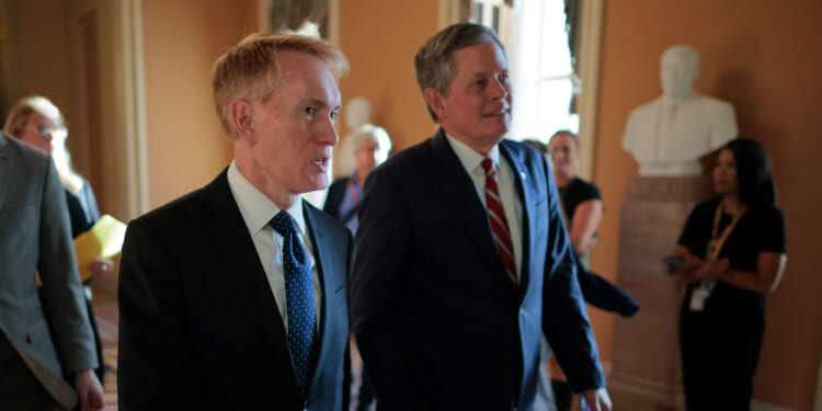 Sen. James Lankford and Sen. Steve Daines in suits walk through a hallway with others in the background. A white statue is visible on a pedestal, and sunlight streams through windows, casting a warm glow on the scene.