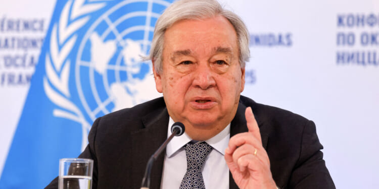 A man in a suit speaks at a podium with a microphone, gesturing with his finger raised. Behind him is a UN logo and a glass of water on the table.