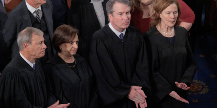 Four Supreme Court justices wearing black robes stand closely together during a formal event, surrounded by other attendees. They appear solemn, with their hands clasped or at their sides.