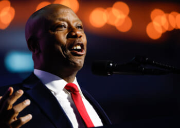 A man in a suit and red tie speaks into a microphone, gesturing with his hand. Warm lights are blurred in the background.