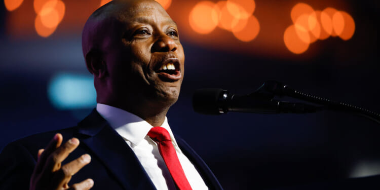 A man in a suit and red tie speaks into a microphone, gesturing with his hand. Warm lights are blurred in the background.