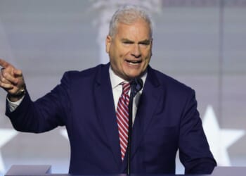 A man in a navy suit and striped tie speaks passionately at a podium, gesturing with his right hand. A blurred background with white star shapes is visible behind him.