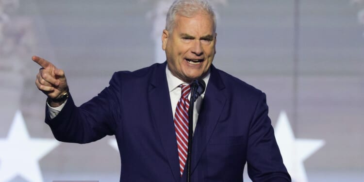 A man in a navy suit and striped tie speaks passionately at a podium, gesturing with his right hand. A blurred background with white star shapes is visible behind him.