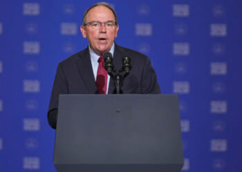 Tom Tiffany in a suit and tie speaks at a podium with multiple microphones, in front of a blue background with repeating white text.