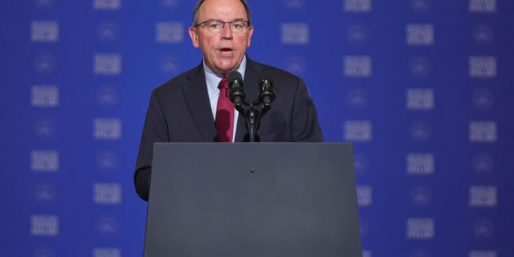 Tom Tiffany in a suit and tie speaks at a podium with multiple microphones, in front of a blue background with repeating white text.