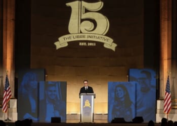 A speaker stands at a podium with The Libre Initiative Est. 2011 projected above. Blue portraits and two American flags flank the stage in a grand hall.