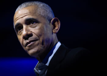 Barack Obama with short gray hair wearing a dark suit and white shirt looks thoughtful against a dark blue background with dramatic lighting.