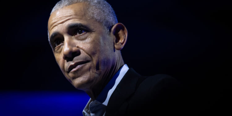 Barack Obama with short gray hair wearing a dark suit and white shirt looks thoughtful against a dark blue background with dramatic lighting.