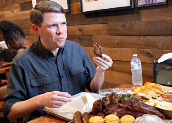 James Talarico sits at a wooden table, eating barbecue with sides like cornbread, greens, and macaroni. He holds a rib. A water bottle, napkins, and sauces are also on the table. Another person sits in the background.