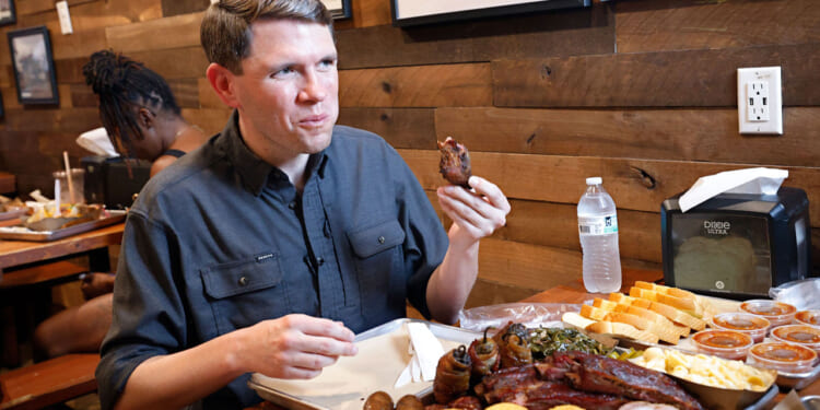 James Talarico sits at a wooden table, eating barbecue with sides like cornbread, greens, and macaroni. He holds a rib. A water bottle, napkins, and sauces are also on the table. Another person sits in the background.