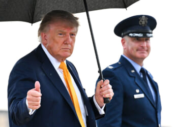 Donald Trump in a suit holds an umbrella and gives a thumbs up, standing next to a uniformed officer outdoors.