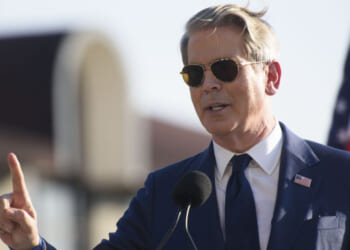 A man in a blue suit and sunglasses speaks at a podium outdoors, gesturing with one finger raised. An American flag is visible in the background.